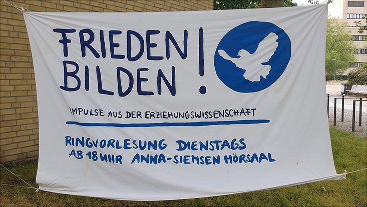 Weißes Banner mit blauer Auschrift "Frieden bilden" hängt zwischen zwei Bäumen.