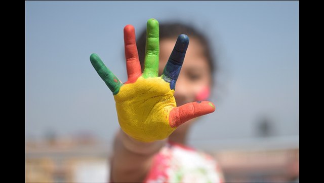 Kind streckt angemalte, bunte Hand nach vorne aus, dem Betrachter entgegen und lacht