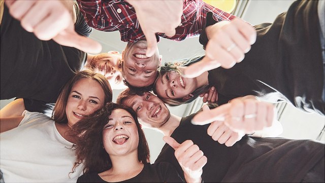 Eine Gruppe steht lachend im Kreis, den Blick nach unten in die Kamera gesenkt